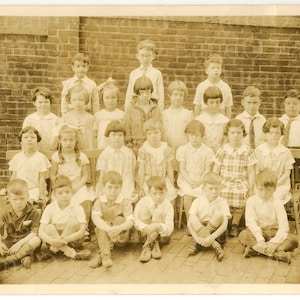 May include: A black and white vintage photograph of a classroom of children, all dressed in clothing from the 1920s. The children are sitting in rows on chairs in front of a brick wall. There are 18 children in the photo, all looking at the camera.