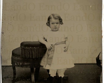 May include: A young girl in a white dress with a red, white, and blue ribbon around her waist sits on a stool with a fringed cushion. She is wearing black shoes and has her hair pulled back.