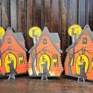 May include: Vintage Halloween house decorations. The orange houses have black roofs and feature a witch with a staff in a yellow window. A black cat is in the window. The houses have a tree and a yellow moon with a lightning bolt.