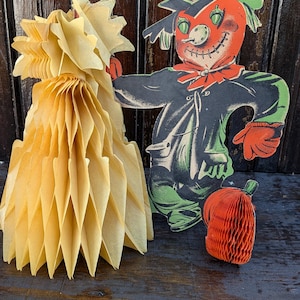 May include: Vintage Halloween decorations including a honeycomb paper pumpkin and a scarecrow figure. The scarecrow has a red face, green eyes, and a black hat and coat. The decorations are on a dark wooden surface.