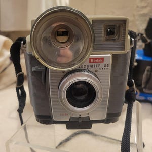 May include: A vintage Kodak Flashmite 20 camera with a black strap. The camera is silver and gray with a black lens and a silver flash attachment. The camera has a dial for color and f-stop settings.