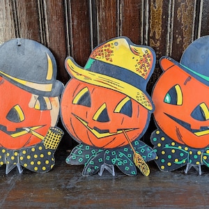 May include: Three vintage Halloween pumpkin decorations. Each pumpkin has a smiling face, a hat, and a decorative collar. The pumpkins are orange, black, yellow, and green. They are made of cardboard and have a small stand.