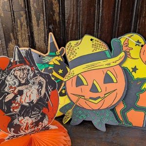 May include: A collection of vintage Halloween decorations, including cutouts of a smiling jack-o'-lantern, a witch, and a black cat. An orange honeycomb paper decoration is in the foreground. The decorations are on a dark wooden surface.