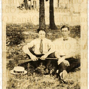 Postal antigua con foto real de padre e hijo relajándose bajo un árbol sosteniendo un rifle tirador de guisantes, década de 1920