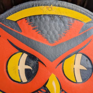 May include: Close-up of a vintage owl design. The owl features a red body with black and yellow accents. The eyes are yellow with a cross-shaped design. The owl is a classic Halloween decoration.
