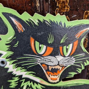 May include: A vintage Halloween decoration depicting a black cat with green and orange details. The cat has a fierce expression with sharp teeth and intense eyes. The cat's fur is black with green edging.