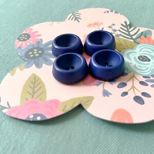 May include: Four dark blue plastic buttons with a raised centre and two holes. The buttons are on a pink background with a floral pattern.