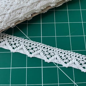 May include: White crochet lace trim with a scalloped edge. The lace is delicate and intricate, with a repeating pattern of small flowers and leaves.