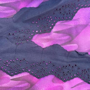 May include: Close-up of navy blue eyelet lace trim with a scalloped edge. The lace features a floral design with small circular cutouts. The trim is set against a vibrant purple and pink floral fabric background.