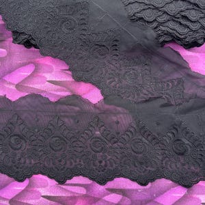May include: Black embroidered lace trim with a scalloped edge. The trim features a detailed floral and scroll design. The lace is layered on a vibrant pink and purple patterned fabric, creating a striking contrast.
