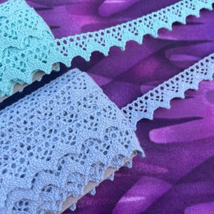 May include: Close-up of two rolls of lace ribbon in shades of blue and teal. The lace has a scalloped edge and an open, airy design. The ribbon is displayed against a purple patterned background.