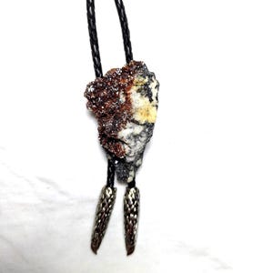 Puede incluir: Una corbata bolo con un colgante de piedra natural en tonos marrón, blanco y amarillo. La piedra cuelga de un cordón trenzado negro, rematado con puntas decorativas plateadas. Este accesorio único añade un toque de elegancia natural.