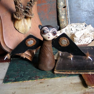 May include: A small, handmade, brown leather bat with black wings and white feathers. The bat has a black and white face with large, round eyes. The bat is sitting on a wooden surface.