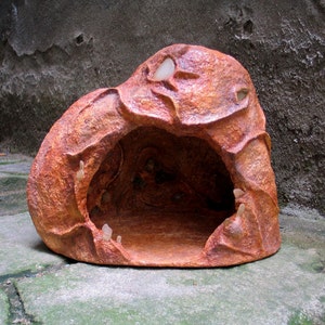 May include: A handmade terracotta cave with a rough, textured surface and a large opening. The cave is painted in shades of brown and orange, with white accents.