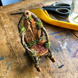 May include: A miniature chair crafted from twigs and moss, featuring a brown and orange fabric seat. The chair is resting on a wooden surface.