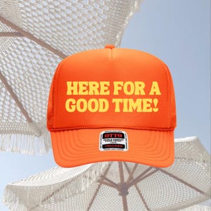 Op de afbeelding: Fel oranje truckerpet met de tekst "HERE FOR A GOOD TIME!" in gele letters. De pet heeft een mesh achterkant en een gebogen klep. Een klein wit label is zichtbaar aan de voorkant.