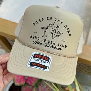 Könnte beinhalten: Beige Trucker-Cap mit Mesh-Rückseite und gebogenem Schirm. Der Hut trägt den Text "TOES IN THE SAND RING ON HER HAND Jenn's Bachelorette" mit Illustrationen. Ein Aufkleber der Marke "OTTO" ist sichtbar.