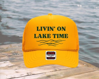 May include: Yellow trucker hat with the text "LIVIN' ON LAKE TIME" in green, along with a wave graphic. The hat has a mesh back and a curved brim. A small label is on the front.