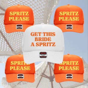 May include: A collection of trucker hats with the text "SPRITZ PLEASE" in orange on an orange cap and "GET THIS BRIDE A SPRITZ" on a white cap. The hats have a mesh back and a curved brim.