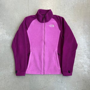 May include: A two-tone fleece jacket featuring a bright pink center panel and deep purple sleeves and collar. The jacket has a full-length front zipper, two zippered side pockets, and the white The North Face logo embroidered on the upper chest.