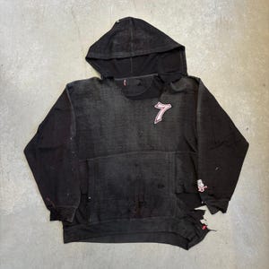 May include: A distressed black hooded sweatshirt featuring a large white and red number 7 patch on the upper chest. The garment has a front kangaroo pocket, long sleeves with ribbed cuffs, and visible fraying and small holes throughout the fabric.
