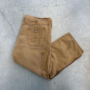 May include: Tan work trousers with multiple pockets, including a large back pocket and a utility pocket on the leg. The trousers have a classic workwear style, with a durable, slightly worn appearance. The Dickies logo is visible on the back pocket.