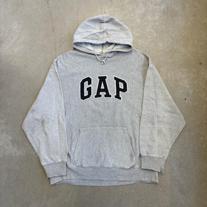 May include: A light grey hooded sweatshirt featuring the word GAP in dark navy blue block letters across the chest. The garment includes a front kangaroo pocket, long sleeves with ribbed cuffs, and a matching grey drawstring at the neckline.