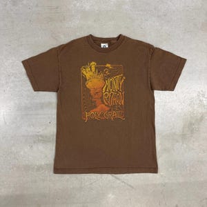 Peut inclure: T-shirt marron avec un graphisme du film "Monty Python et le Saint Graal". Le motif, en orange et jaune, comprend le titre du film et une image stylisée d'une couronne. Le t-shirt a un col rond et des manches courtes.