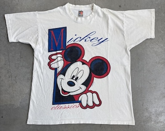 May include: White t-shirt featuring a graphic of Mickey Mouse. The design includes Mickey's face and hands, with the word "Mickey" in blue script and the word "classics" in red. The graphic is framed by a blue and red border.
