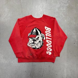 May include: Red crewneck sweatshirt featuring a bulldog graphic wearing a black and white hat with a "G" on it. The word "BULLDOGS" is printed vertically in white on the right side of the sweatshirt.