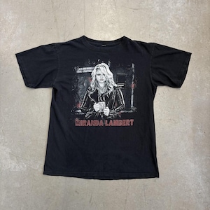 May include: A black short-sleeve t-shirt featuring a graphic print of a person with long blonde hair wearing a leather jacket. The print includes a distressed background and the text MIRANDA LAMBERT in red capital letters at the bottom.