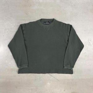 Peut inclure: Un t-shirt thermique à manches longues vert olive foncé. Le t-shirt a une texture côtelée et un col ras du cou avec un petit détail cousu. Le logo de la marque est visible en haut à gauche. Les poignets et l'ourlet montrent des signes d'usure.