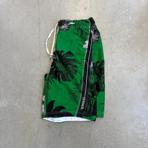 May include: Green swim trunks featuring a black tropical leaf pattern and a white drawstring at the waist. The shorts include a vertical black side panel with the white Speedo logo printed along the seam, and a white interior lining visible at the hem.