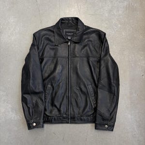 May include: A black leather jacket featuring a full-length front zip, a classic pointed collar, and long sleeves with buttoned cuffs. The garment includes two side pockets and a visible Van Heusen brand label stitched inside the neckline.
