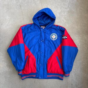 Könnte beinhalten: Eine Kapuzenjacke in auffälligem Colorblock-Design. Die Jacke hat eine lebendige Kombination aus roten und blauen Paneelen, einen runden Aufnäher auf der Brust und einen "7"-Aufnäher auf dem Ärmel. Sie hat eine Knopfleiste und einen gerippten Saum.