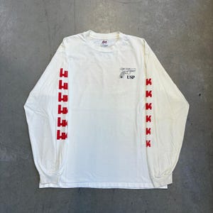 Peut inclure: Un t-shirt blanc à manches longues avec un petit graphique noir représentant une arme de poing et le texte "USP" sur la poitrine droite. Les deux manches sont ornées d'un motif vertical répétitif de la lettre "K" rouge en police grasse.
