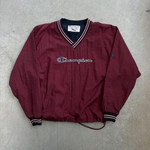 May include: A maroon Champion pullover windbreaker with a V-neck collar. The collar and cuffs have white and navy blue stripes. The Champion logo is embroidered across the chest in white script. A small Champion logo patch is on the right sleeve.