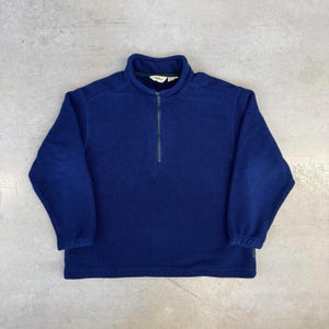 May include: A navy blue fleece pullover with a half-zip closure and a stand-up collar. The zipper is green. The long-sleeved top is made of a soft, warm material. The garment is laid flat on a gray surface.
