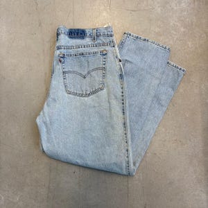 May include: Light blue acid-wash denim jeans featuring a high-waisted design, a button fly closure, and two back pockets with a signature arcuate stitch pattern. The jeans have a straight-leg cut and include a small red tab on the back pocket.