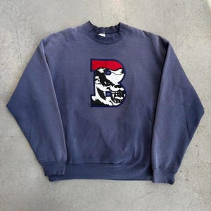May include: A faded navy blue sweatshirt with a crew neck. The sweatshirt features a large, embroidered letter "B" with a red top and a black and white bulldog graphic inside. The sweatshirt has a vintage, worn look.