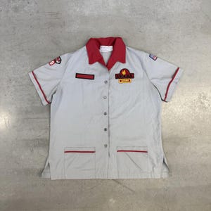 May include: A light grey short-sleeved button-down shirt with a red collar and red trim on the sleeves and pockets. The shirt features an Awana Leader patch on the chest, a small American flag patch on the sleeve, and a rectangular name tag space.