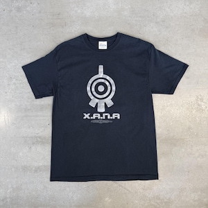 May include: A black short-sleeved t-shirt featuring a large, silver, circular graphic symbol centered on the chest. Below the symbol, the text X.A.N.A is printed in a bold, silver, sans-serif font, with a smaller decorative emblem underneath.