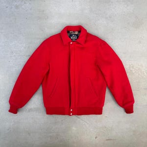 May include: A vibrant red wool jacket featuring a collar, two front pockets, and button fastenings. The jacket has ribbed cuffs and hem. The interior collar lining displays a patterned design. The Woolrich brand label is visible.