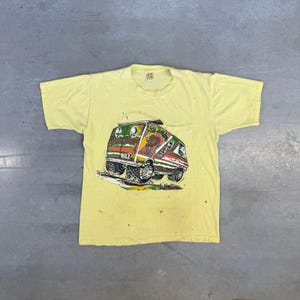 May include: A pale yellow short-sleeve t-shirt featuring a graphic print of a custom van with oversized tyres and the text Hang Ten. The van design includes green, white, and red horizontal stripes, a brown circular logo, and a small chest pocket.