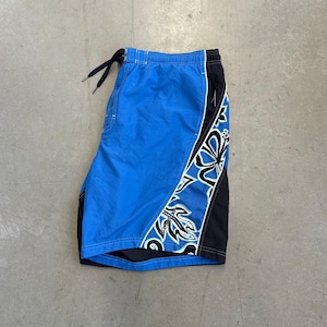 May include: Blue swim trunks featuring a black side panel with a white floral graphic print. The shorts have an elastic waistband with a black drawstring cord and a small white Speedo logo printed on the lower right side of the patterned panel.