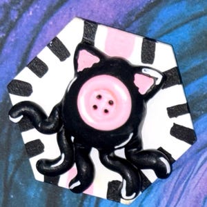 May include: A whimsical, handmade button featuring a black cat-like creature with pink accents and a pink button eye. The creature has tentacles and is set against a black and white striped background.