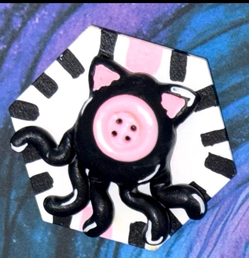 May include: A whimsical, handmade button featuring a black cat-like creature with pink accents and a pink button eye. The creature has tentacles and is set against a black and white striped background.