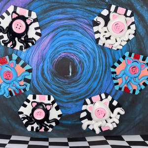 May include: A collection of whimsical, handmade octopus-pig hybrid art pieces. Each piece features a pink button nose, a button eye, and tentacles. The octopuses are in black, white, and blue, set against a hexagonal background with a striped border.
