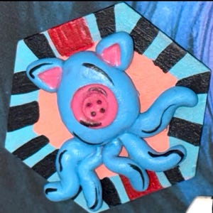 May include: A whimsical, blue octopus-like creature with pink accents and a button-like face. It is set against a colorful, geometric background with red, black, and blue stripes, creating a playful, artistic design.