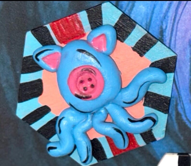 May include: A whimsical, blue octopus-like creature with pink accents and a button-like face. It is set against a colorful, geometric background with red, black, and blue stripes, creating a playful, artistic design.
